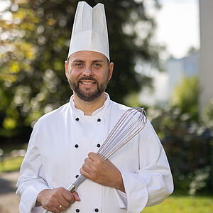Foto eines Mannes in weißer Küchenkleidung mit Küchenhaube und mit großem Schneebesen in der Hand