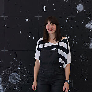 Foto von Marianne Lang vor einem Bild mit einem Sternenhimmel und größeren und kleineren Asteroiden