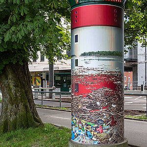 Litfaßsäule mit dem Kunstplakat im Hintergrund die Straßenunterführung des Haus der Natur