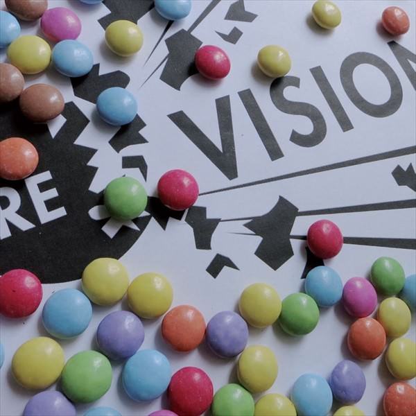 "Re-Vision" Schriftzug "RE - VISION" in schwarz-weiß und darauf verteilt liegend viele bunte Smarties