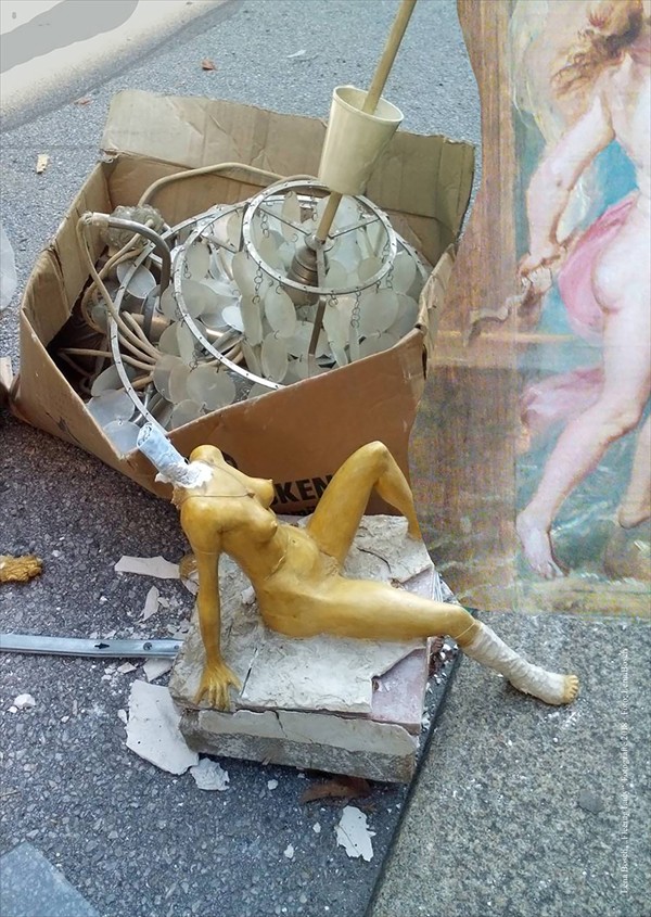 Foto von am Straßenrand abgelegten Dingen: im Vordergrund die Plastik eines nackten Frauenkörpers in sitzender Position, ohne Kopf, dahinter ein großer Lampenschirm aus drei unterschiedlich großen Metallringen, an denen runde Kunststoffkreise an kleinen Ketten hängen in einem Karton.