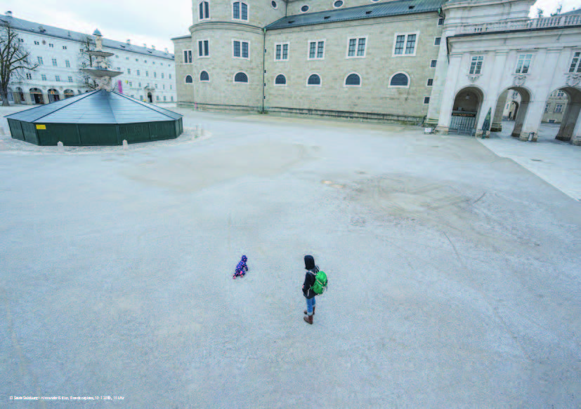 Residenzplatz, 30.3.2020, 15 Uhr Foto vom Residenzplatz mit dem Residenzbrunnen, der mit der Winterverkleidung aus Holz abgedeckt ist. Im Vorderfgrund eine erwachsene Person mit einem spielenden Kind, sonst ist der Platz Menschenleer.