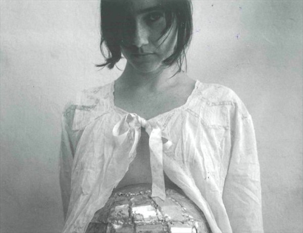 Die zeitgenössische kubanische Fotografie Oberkörperpoträt einer Frau mit dunklem halblangem Haar, gekleidet in eine weiße langärmelige Spitzenbluse die im Ausschnitt mit einer Masche gebunden ist, aus der geöffneten Bluse ist eine glänzende Kugel - in Annäherung einer Weltkugel - ansatzweise zu sehen.