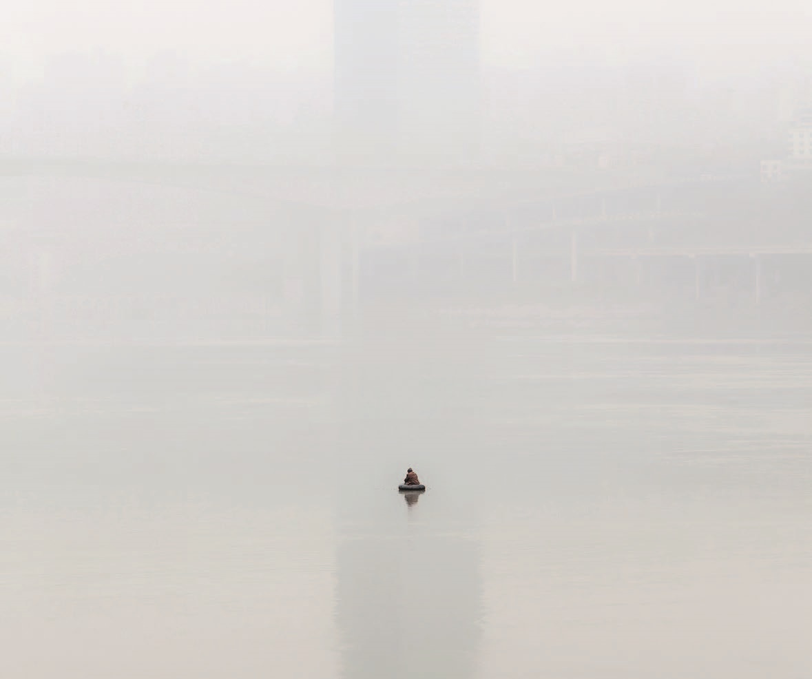 Downstream the Greyscale Nebelverhangenes Bild mit einer Person auf dem Wasser in einem sehr kleinen runden Schlauchboot sitzend. Im Hintergrund im Nebel kaum erkennbare Gebäudestrukturen.