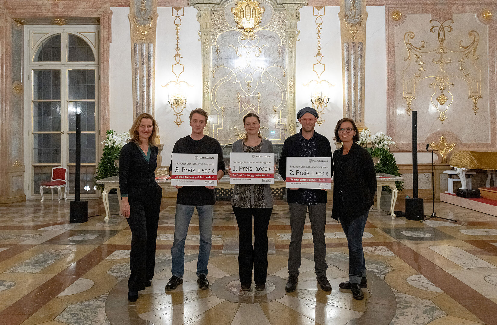 Preisträger*innen des Drehbuchentwicklungspreise 2021 Gruppenfoto im Marmorsaal Schloß Mirabell; die Preisträger halten ihre Preise in Form von übergroßen Gutscheinen vor dem Körper in Händen.